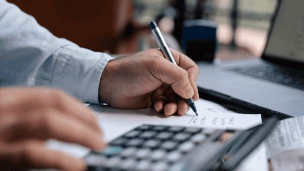 Man writing numbers at desk in front of calculator and laptop