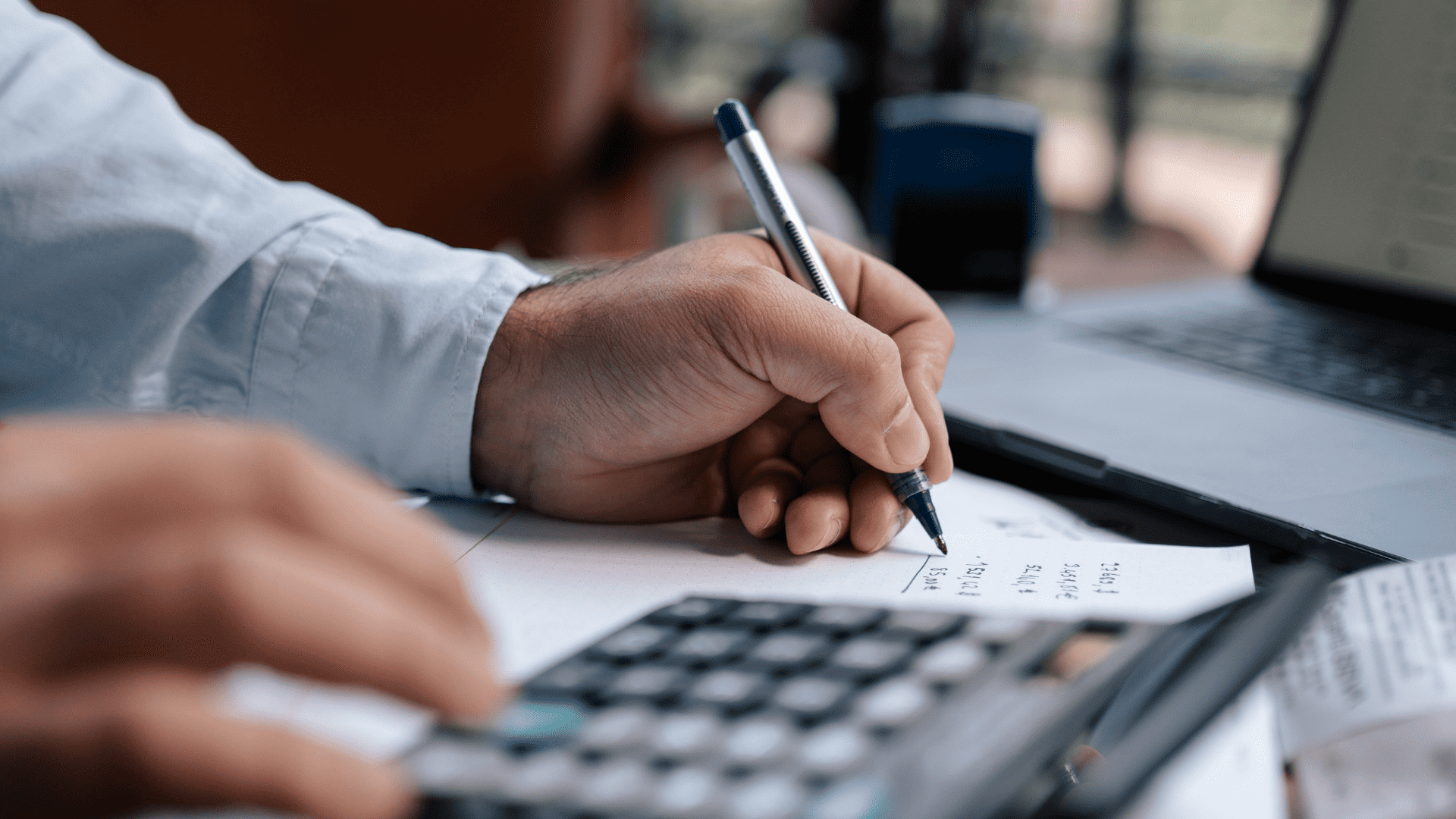 Man writing numbers at desk in front of calculator and laptop